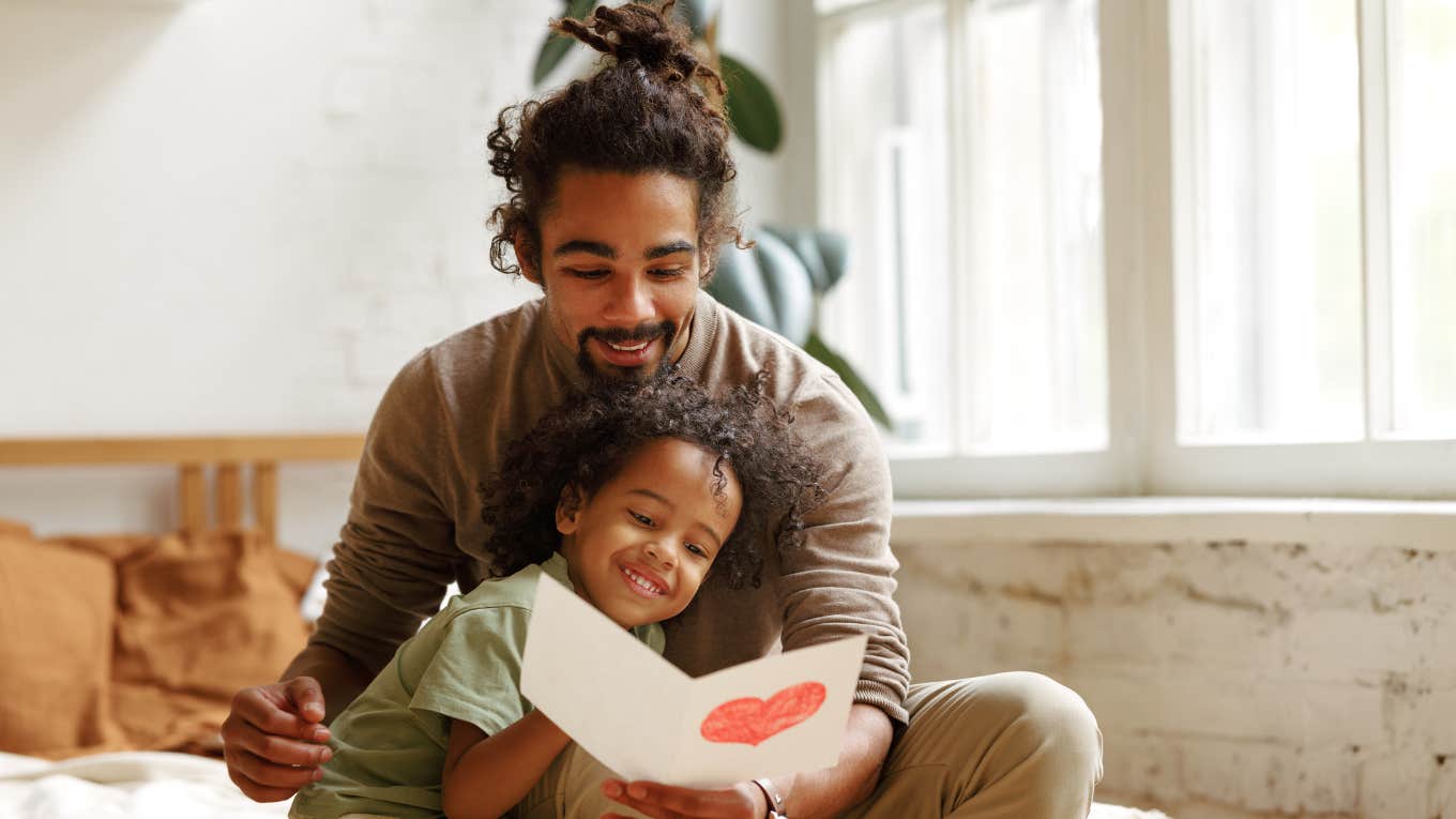 father reading hand-drawn card with young son in bedroom while sitting