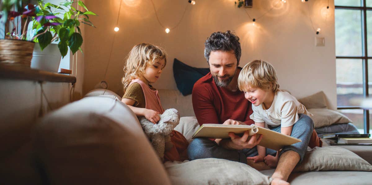 Dad Won't Let His Wife FaceTime Their Kids Anymore After She Moved to NYC Father sitting with two young children on a sofa indoors and flipping through a book.