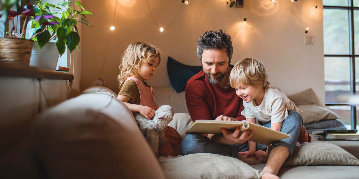 Father sitting with two young children on a sofa indoors and flipping through a book.