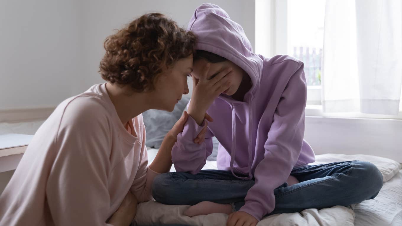Worried mom comforting crying teen daughter at home while sitting on bed