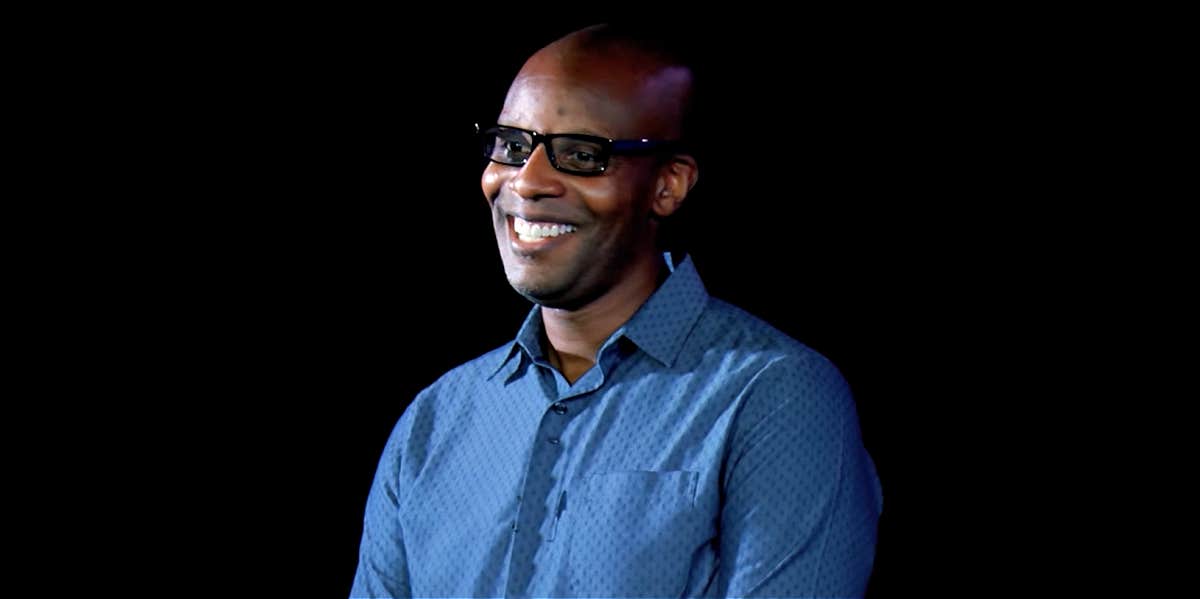 Doyin Richards, a Black woman with a bald head and black glasses, wears a blue shirt and delivers a speech against a dark background, smiling