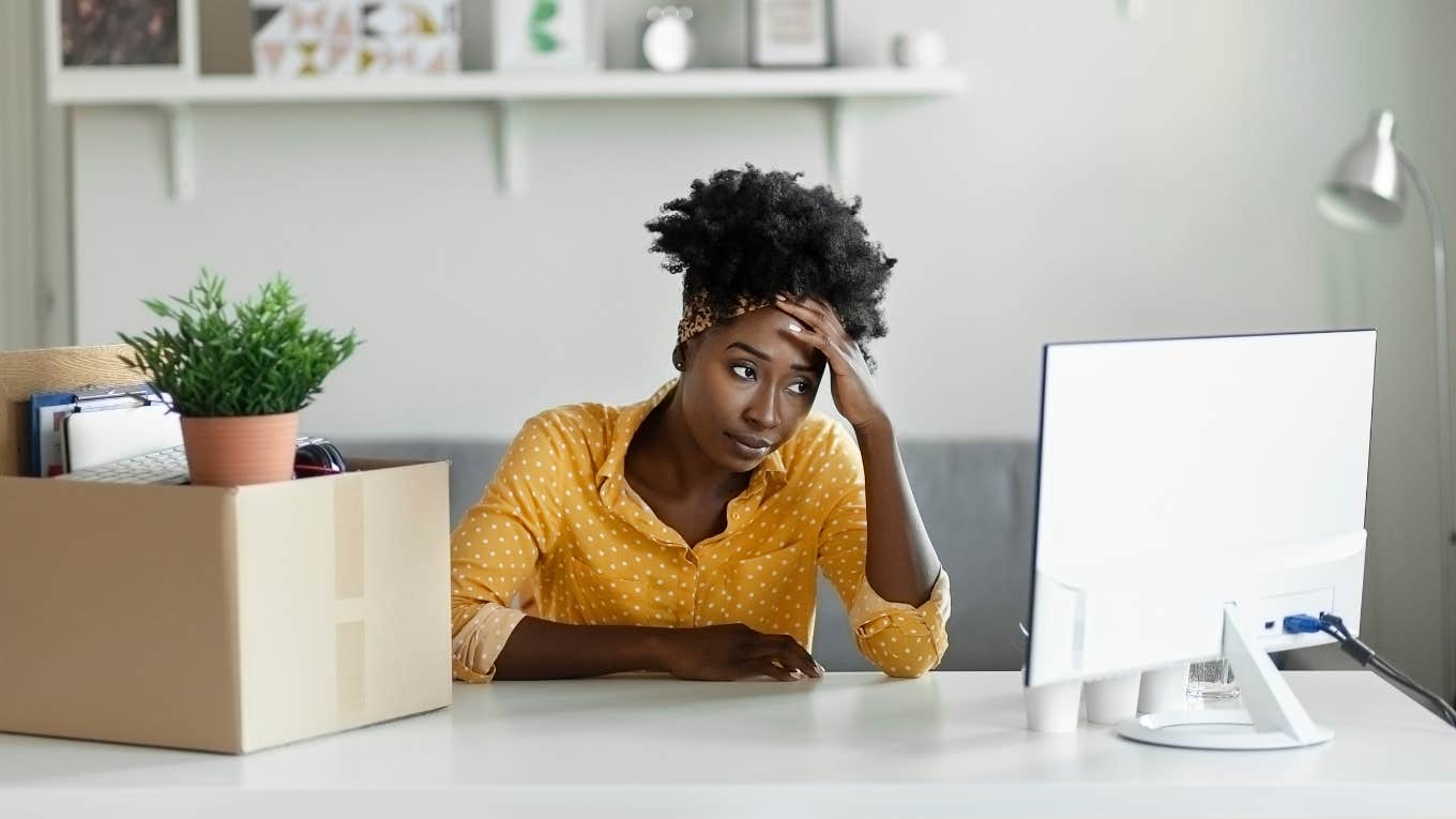 office employee holding hand against forehead after being fired with cardboard box on desk and looking at computer