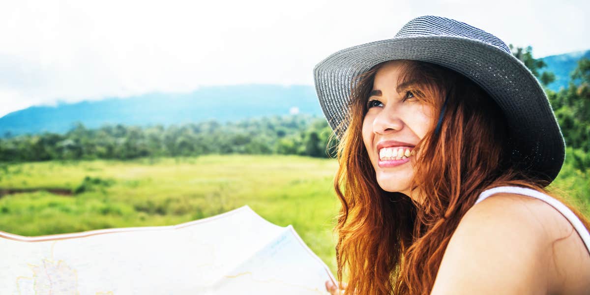 Joyful Asian woman wearing a hat, laughing with a map by a field