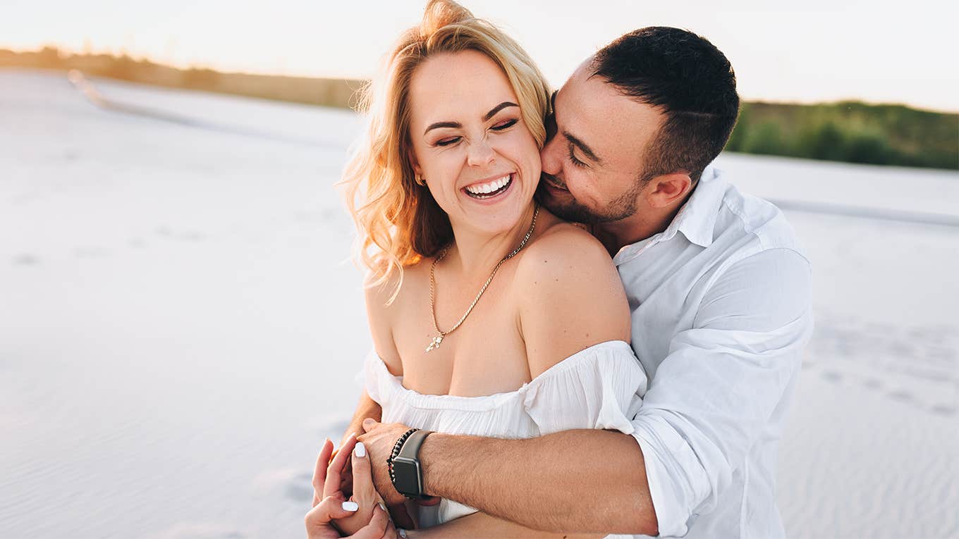 A bearded man and a blond woman hugging against the background of white sand. 