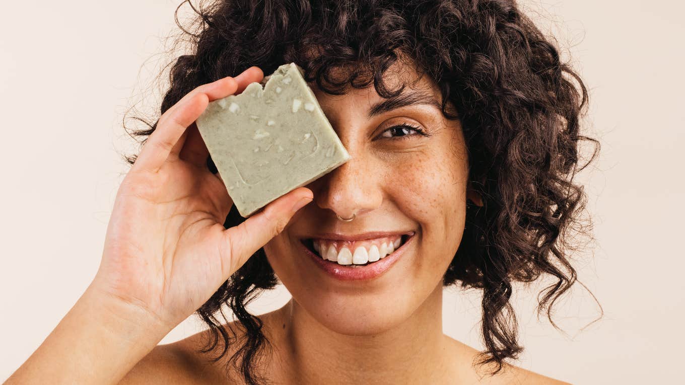young woman smiling at the camera while holding a bar of natural soap close to her face.