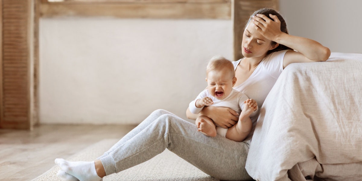Tired infant baby cries in mother hands, depressed unhappy exhausted mom sitting on floor with crying little child on her lap, bedroom interior, copy space. Postnatal postpartum depression concept Tired mom holding crying baby.