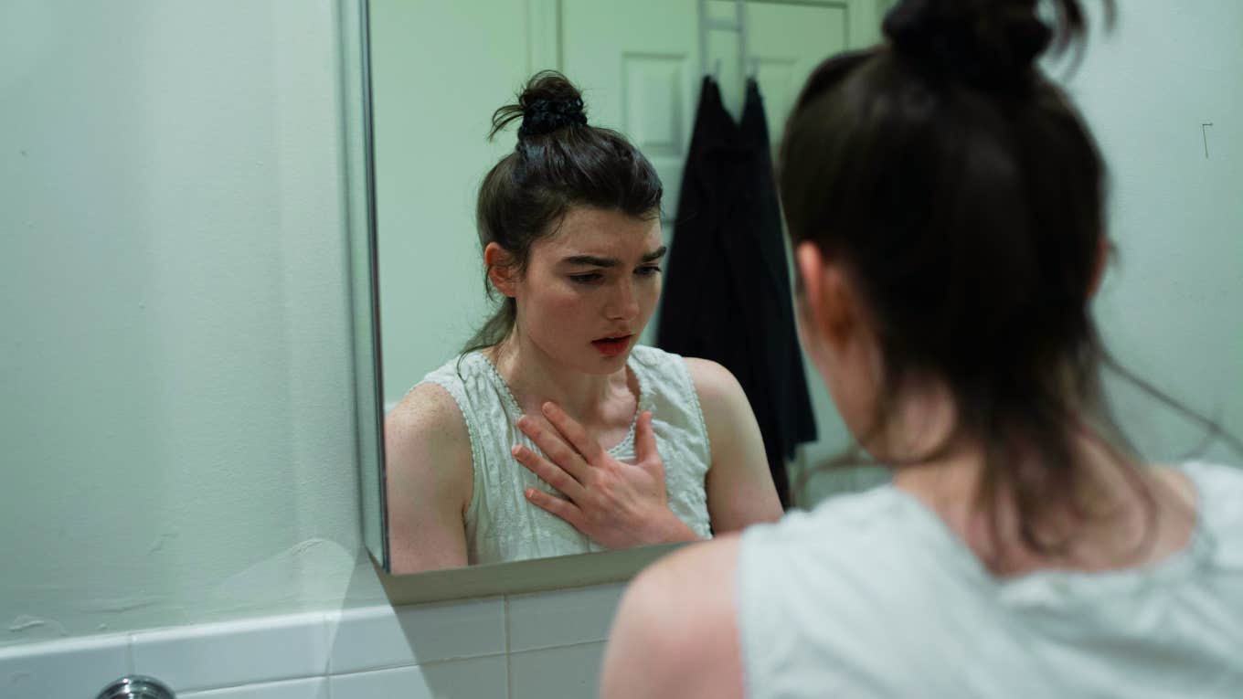 Fundamental Ways Being Cheated On Changes You Disassociated woman in bathroom with hand on chest, unable to look at herself after being cheated on