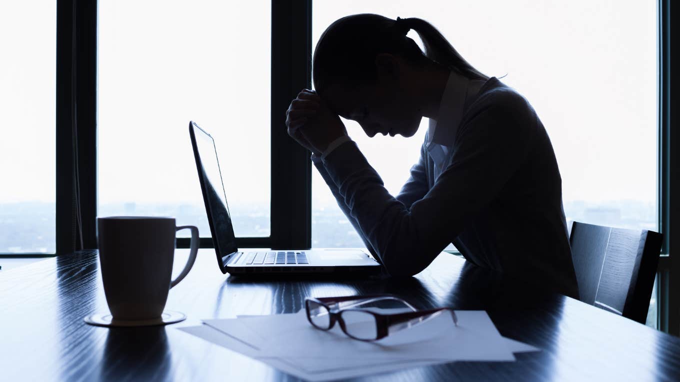 Worker Forced To Return To Work And Smile Despite The Recent Loss Of A Family Member Silhouette of stressed businesswoman with head in hands sitting at desk in front of window