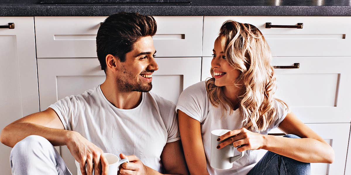 smiling couple sits against kitchen counters drinking coffee