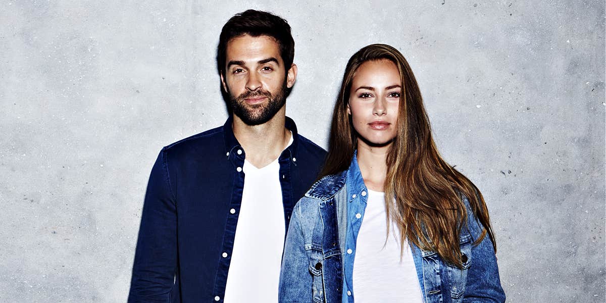 couple standing against a grey wall, both wearing denim jackets and white tees