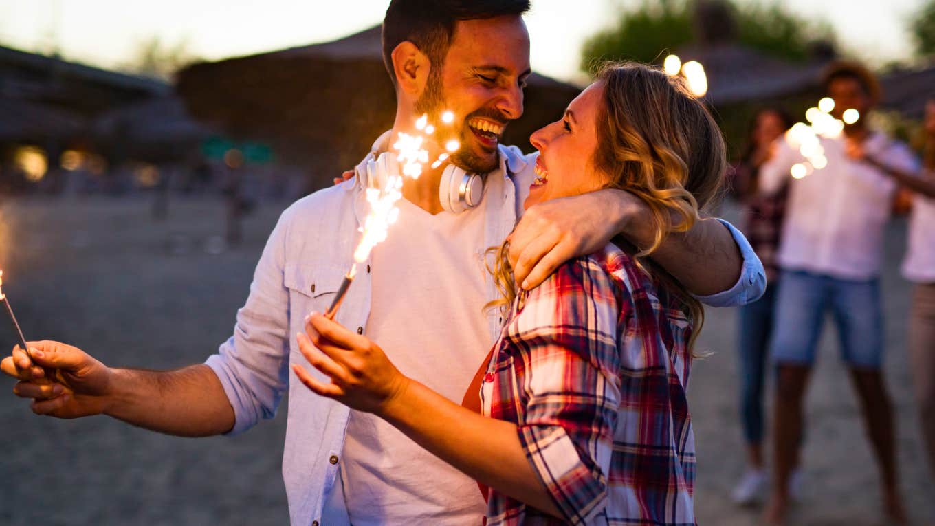 happy couple lighting sparklers at beach during sunset