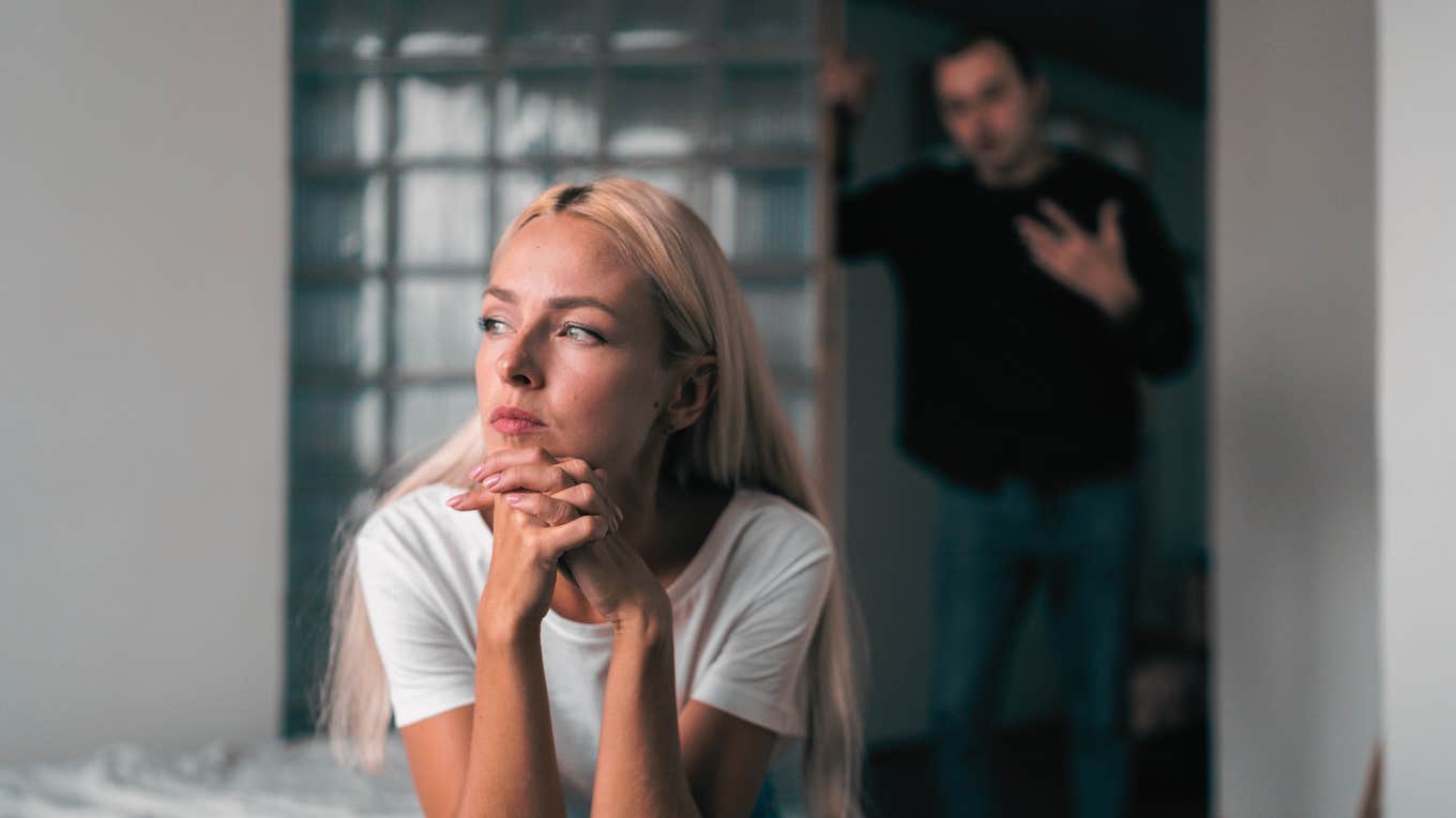 woman looking away sitting on bed husband blurred in background