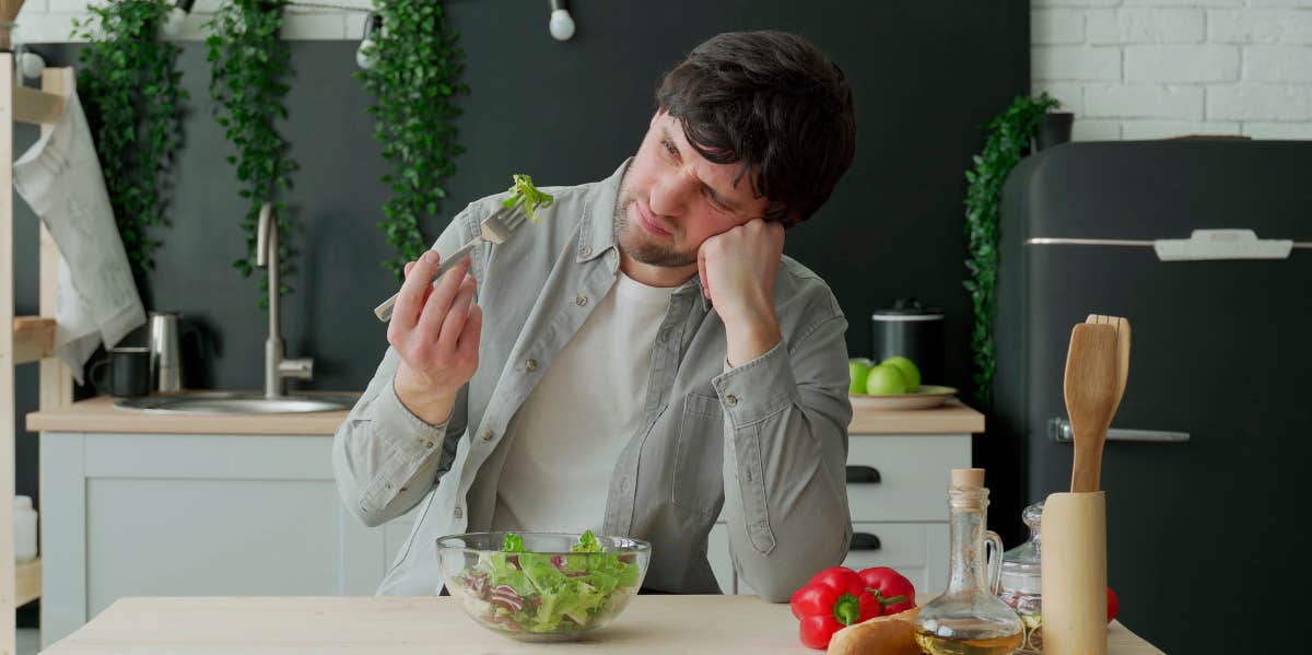 Unhappy man eating vegetable salad at table in kitchen.