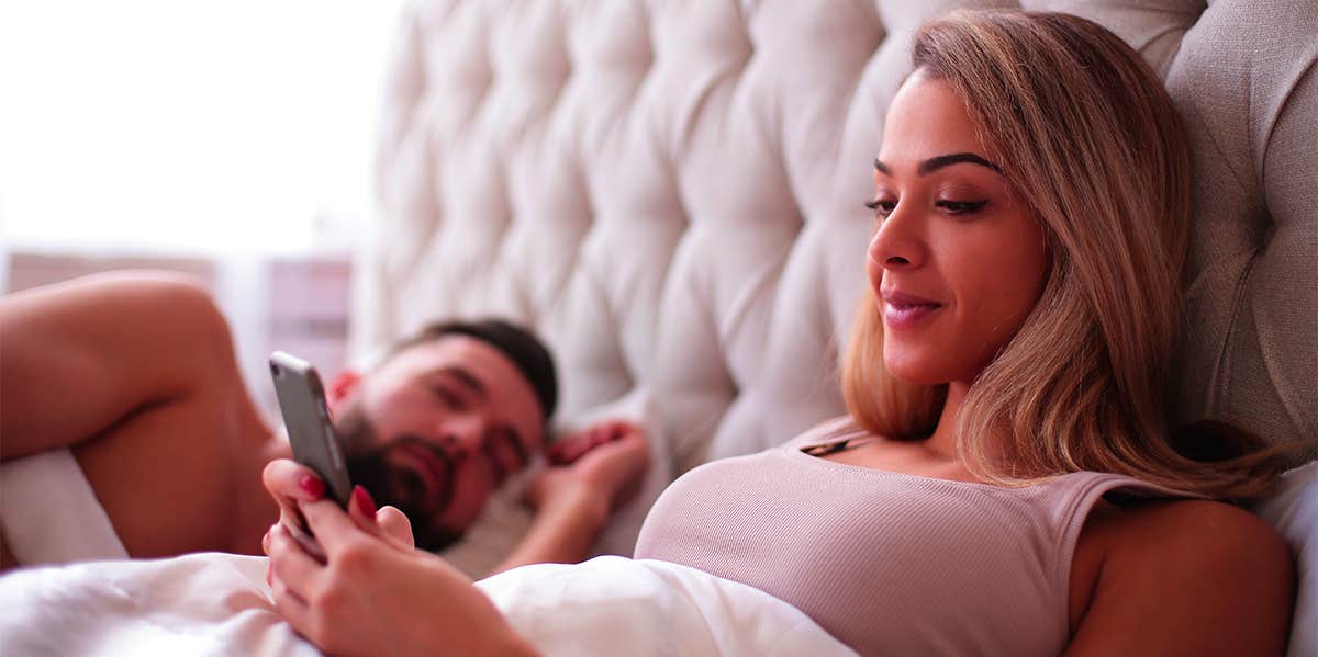 woman laying in bed looking at phone with man sleeping next to her