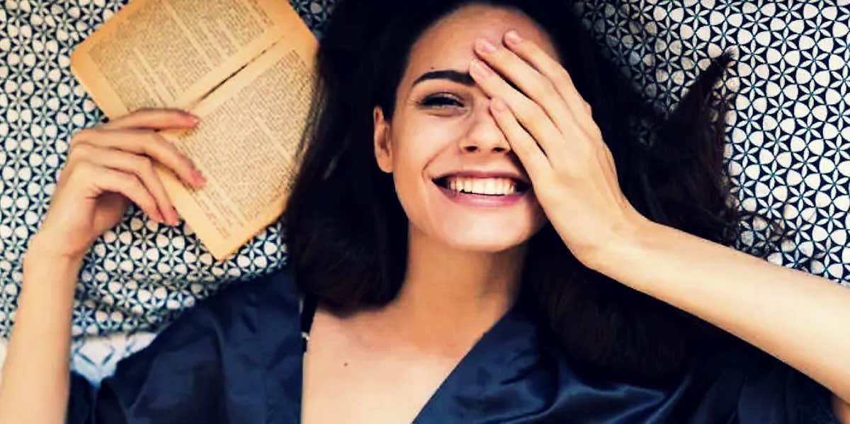 brunette woman smiling, hand over one eye, book in hand on a bed