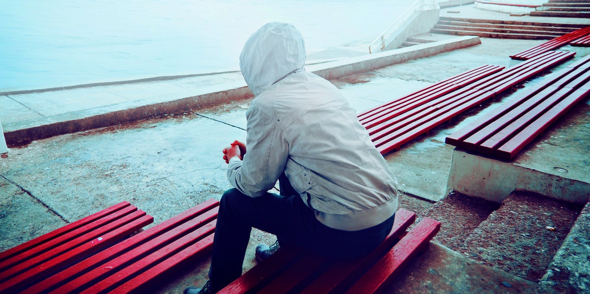 man in hoodie sitting, depressed, on a park bench looking at ocean