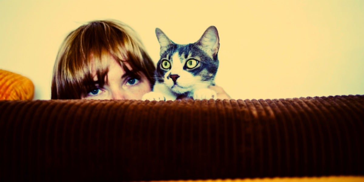 woman with long blonde bangs hides behind couch with her cat