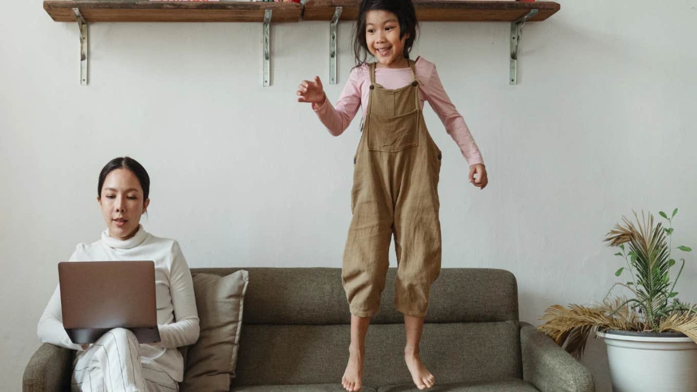 Introverted Mom With Extroverted Kid Wonders If She's A Terrible Parent For Being Overstimulated & Snapping At Her Kid mom on computer while kid jumps on couch
