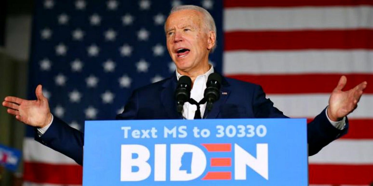 Joe Biden stands at a podium with a Biden campaign sign on it, arms out, speaking, with an American flag behind him
