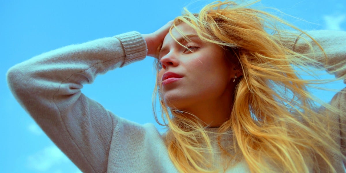 blonde woman with lipstick on, hands in her hair, against blue sky 