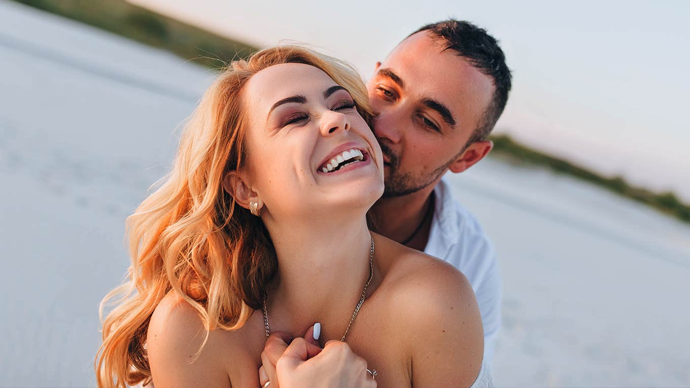 A tanned young caucasian bearded guy kisses a beautiful fun smiling blonde woman against the backdrop of the setting sun.