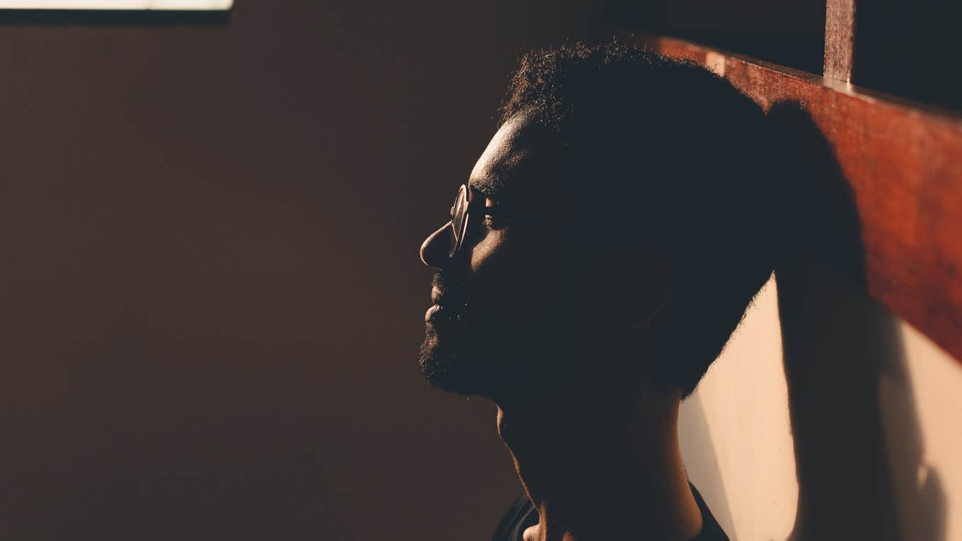 dark side shot of upset young man with glasses sitting alone with back against a wall