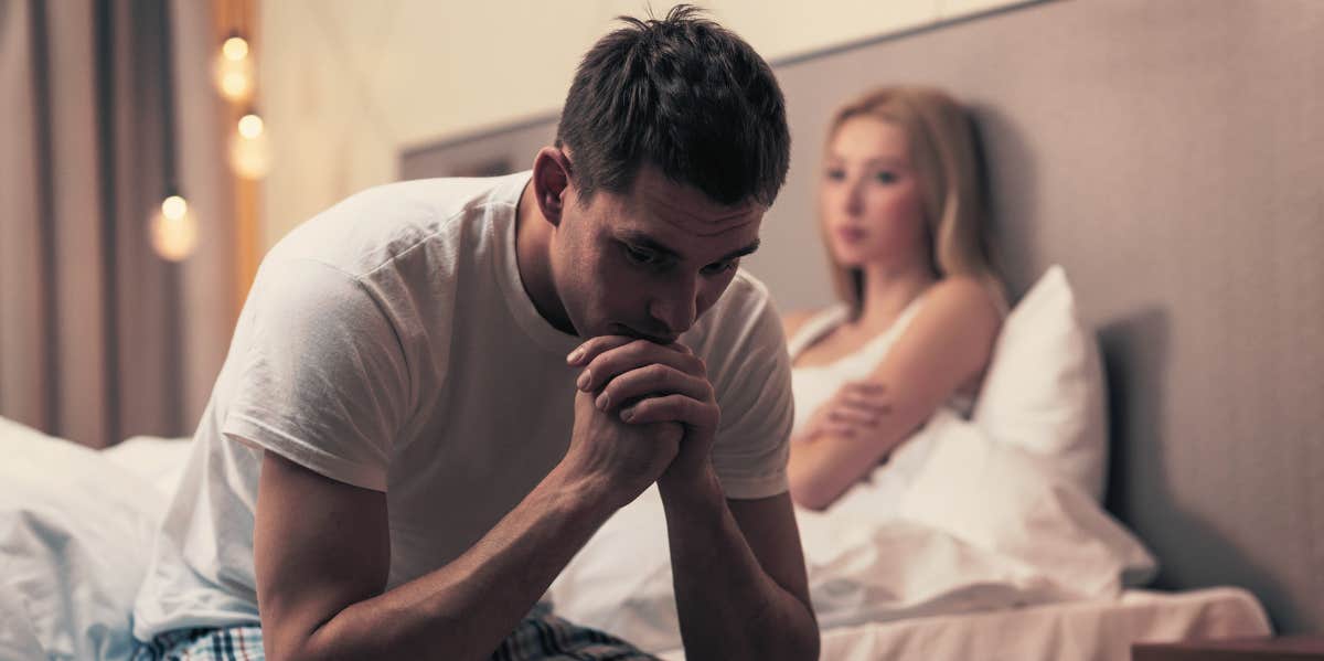 Upset man sitting on the bed with woman in the back