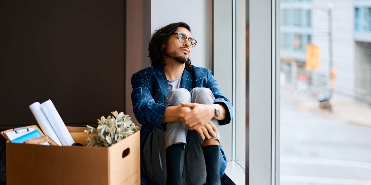 Distressed worker sits by a window with a box of his office things after losing their job.