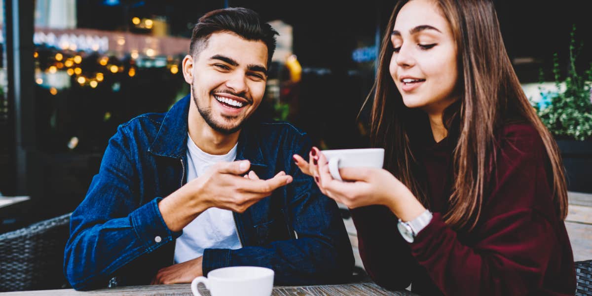 Young adult Latinx couple smiling at a cafe, man gesturing toward the woman who is holding a coffee cup
