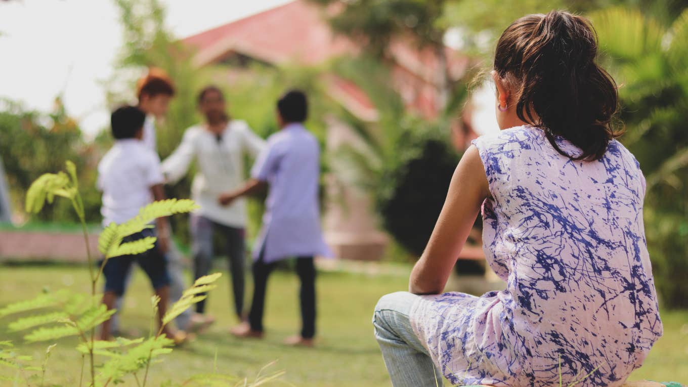 back view of sad little girl sitting alone at park looking at other kids playing