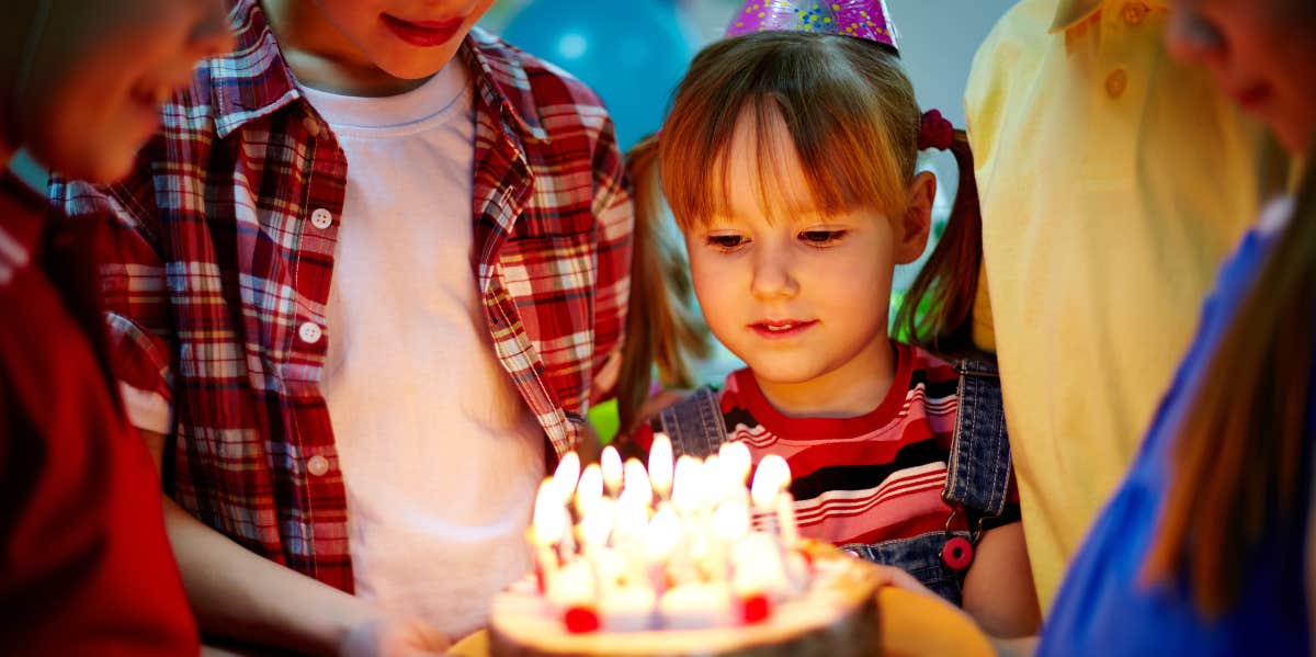 A little girl surrounded by other kids holds a candle-lit cake for her birthday