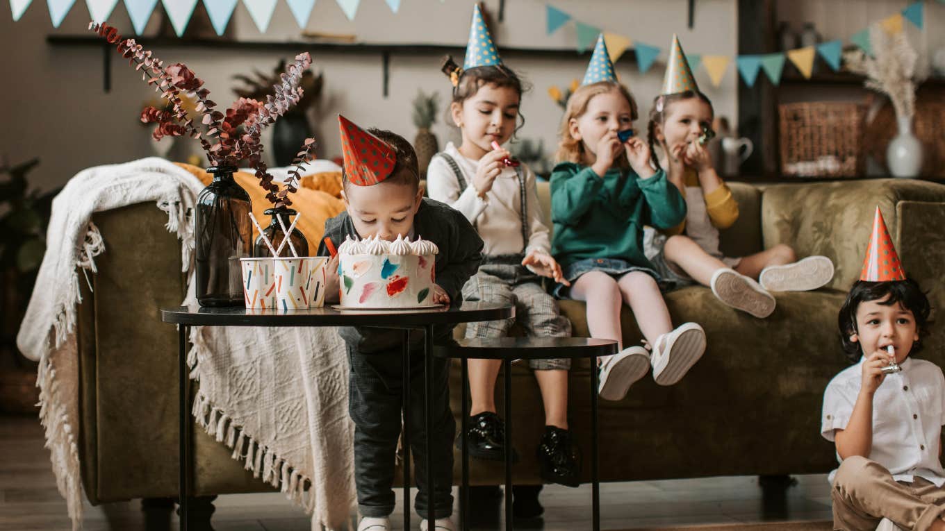 little boy biting into birthday cake while children sit behind him at birthday party
