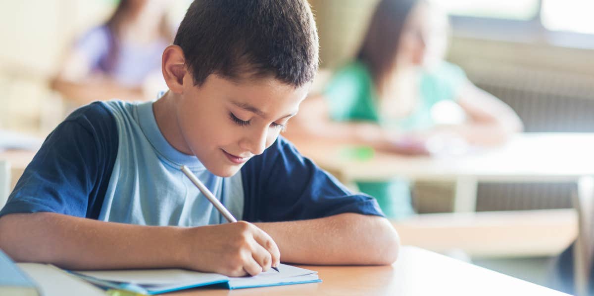 Mom Says Her Son Was In Tears After Reading His Teacher's Note About Test Scores little boy smiling while taking notes in class