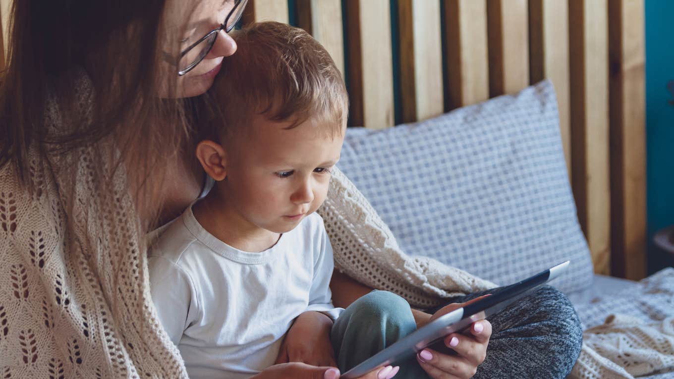 Mom Decides To Try Low Demand Parenting By Letting Her Homeschooled Kid Make His Own Rules & Be On His iPad Most Of The Day mom holding son while he plays on a tablet