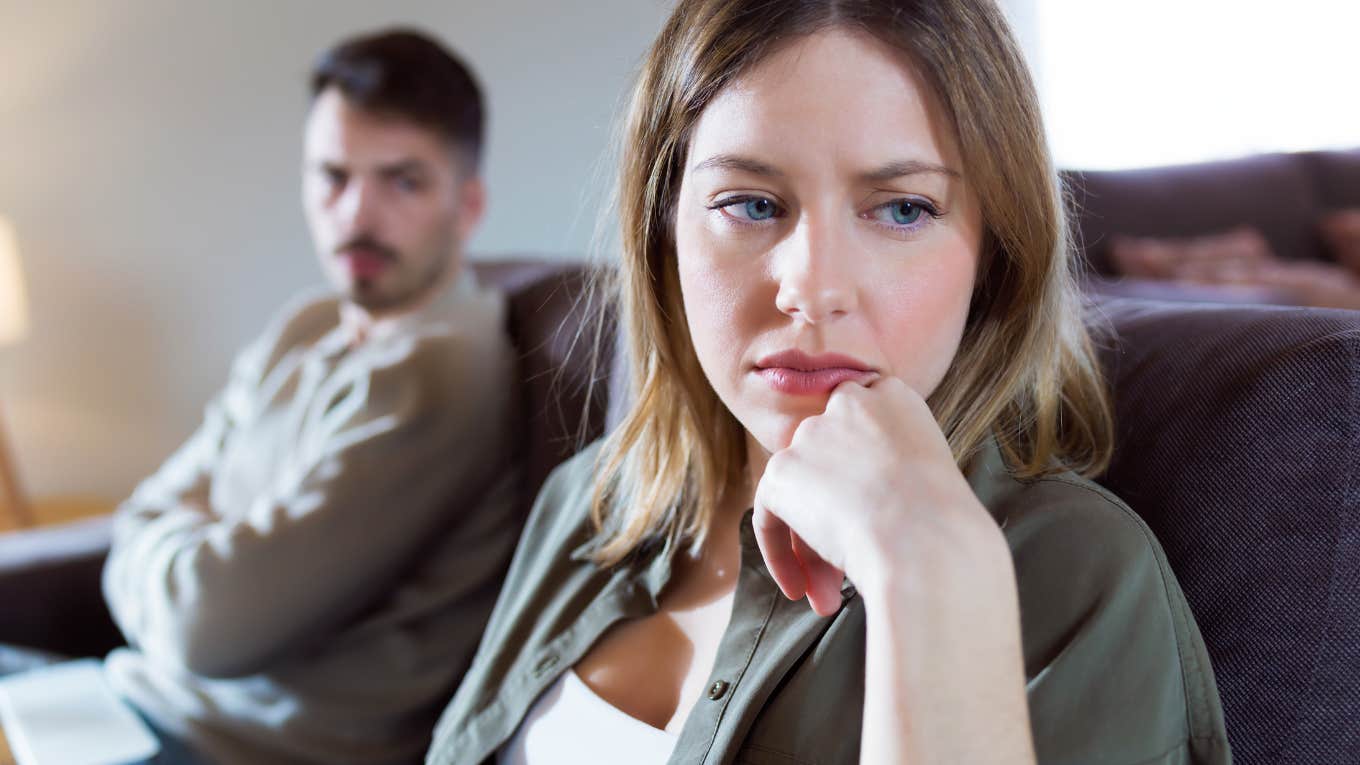 Portrait of offended young woman ignoring her angry partner sitting behind her on the couch at home.