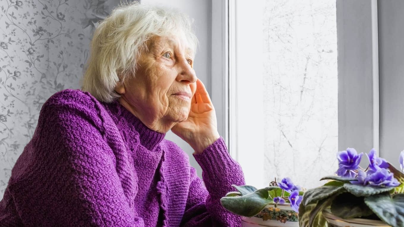 Older woman looks sad while looking out of a window. 