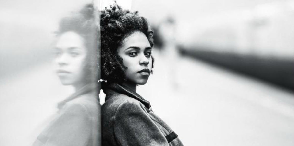 woman with brown skin and curly dark hair leans against a wall, looking sideways at camera