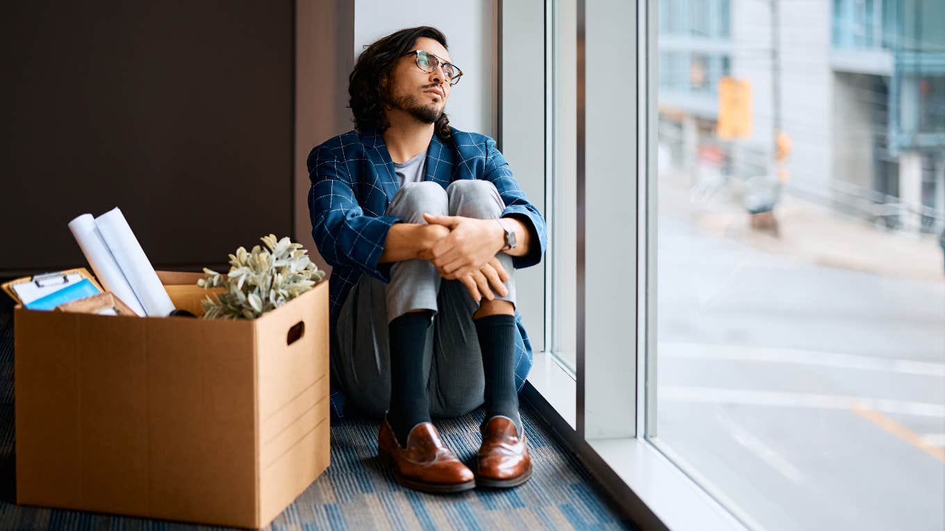 sad employee sitting next to box of his belongings and looking through the window after being fired at work.