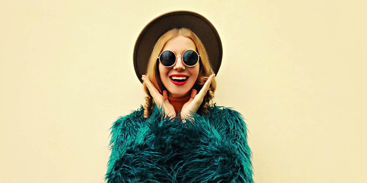 woman wearing a black hat and round glasses, smiling in front of yellow wall