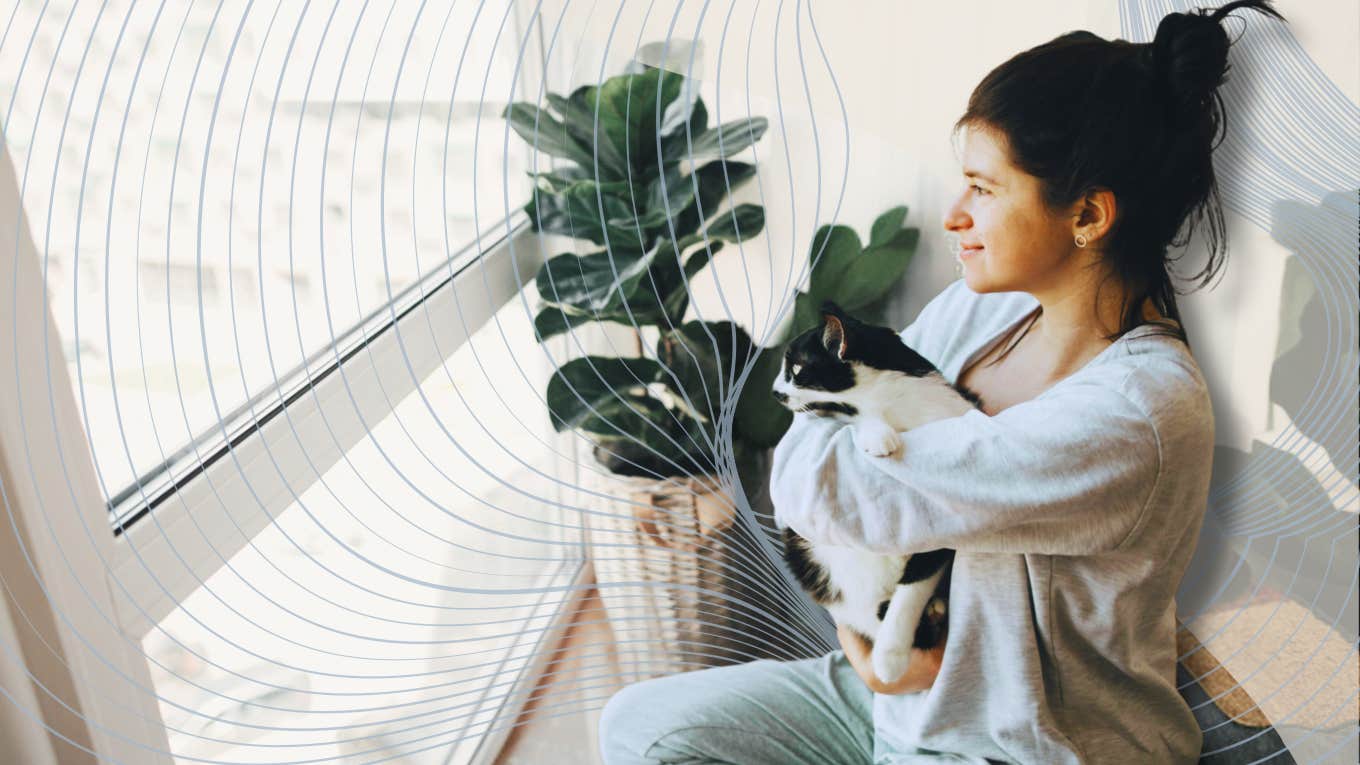 Sensitive woman holding her cat looking out window 