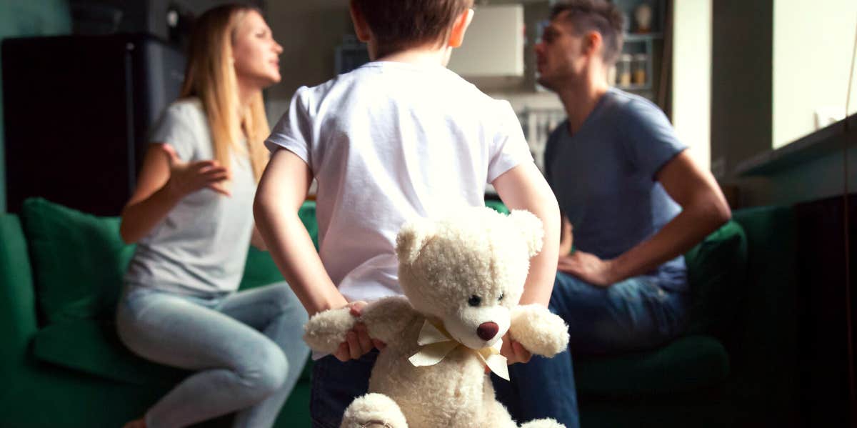 parents sitting on couch fighting in front of little boy holding a teddy bear