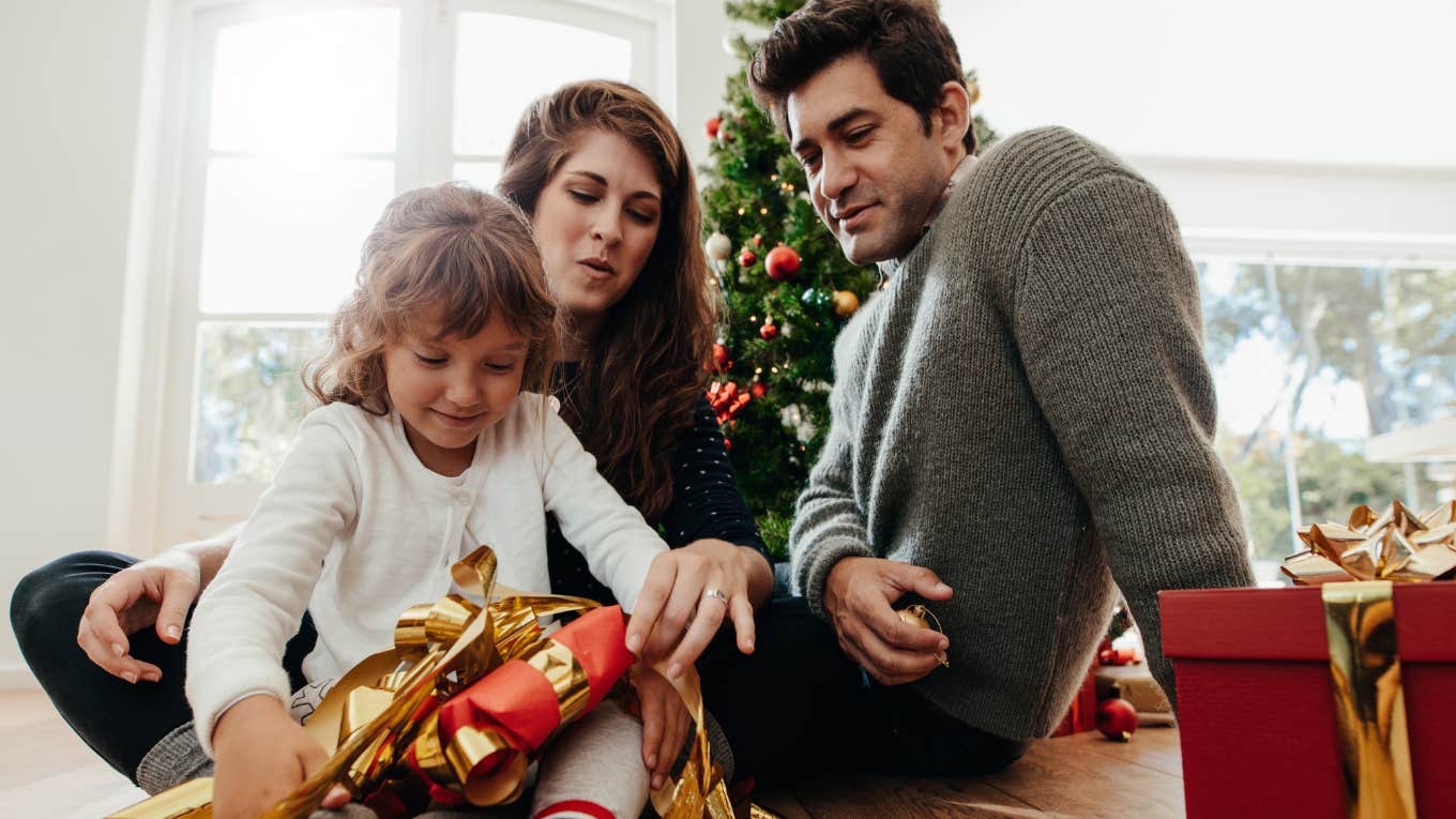 Parents watching their daughter open a Christmas gift. 