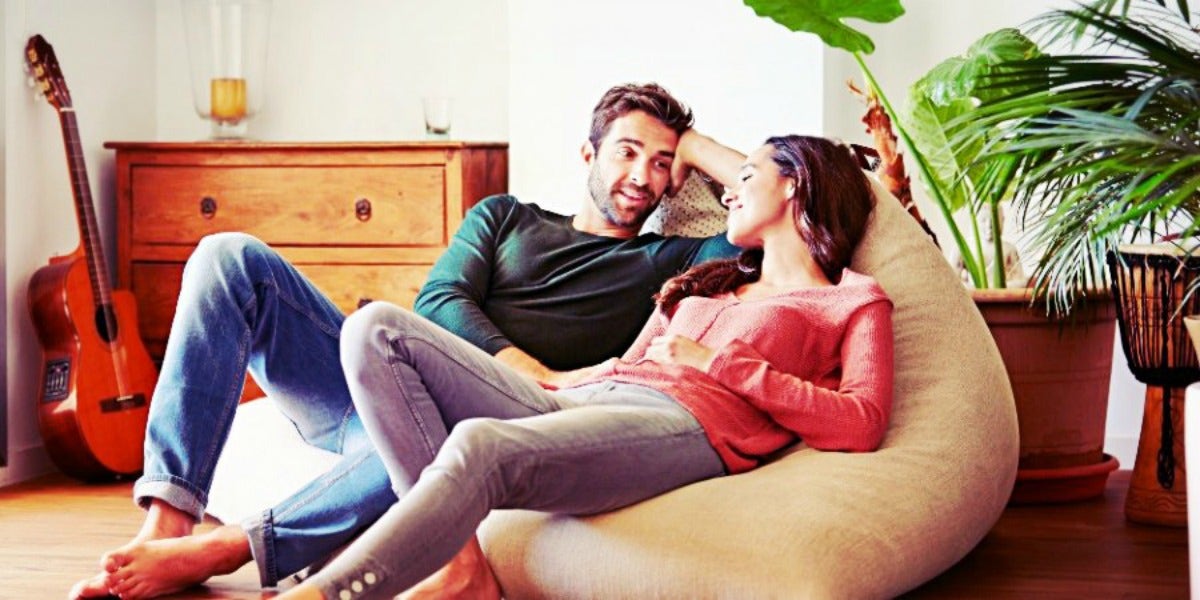 couple lounges in the sunlight, resting on a large pouf chair, looking at one another