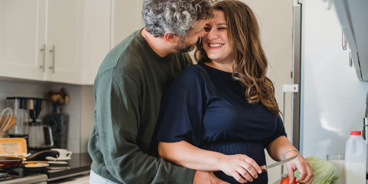Man Makes Pregnant Wife Cry By Calling Her 'Less Of A Person' After She Refuses To Cut Contact With Her Dad husband embracing pregnant wife while standing in the kitchen