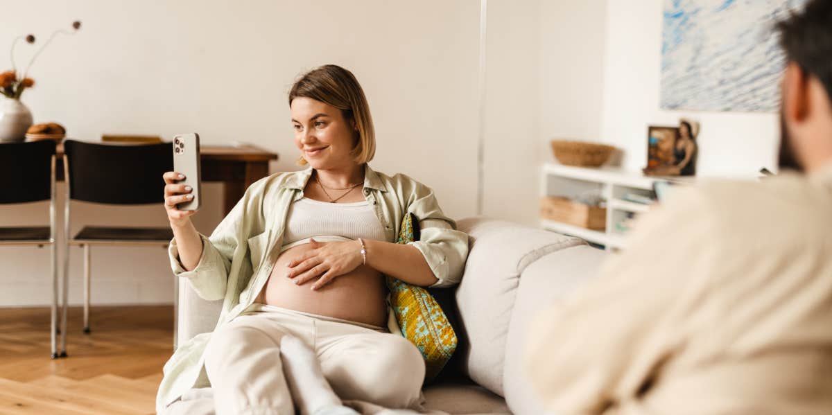 pregnant woman taking selfie on cellphone while sitting at couch in home