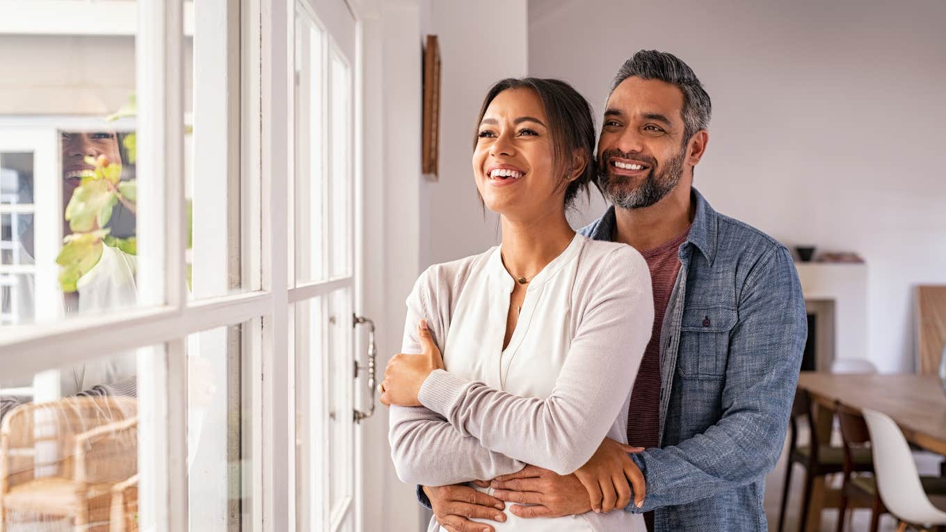 Smiling adult couple hugging each other and standing near window while looking outside.