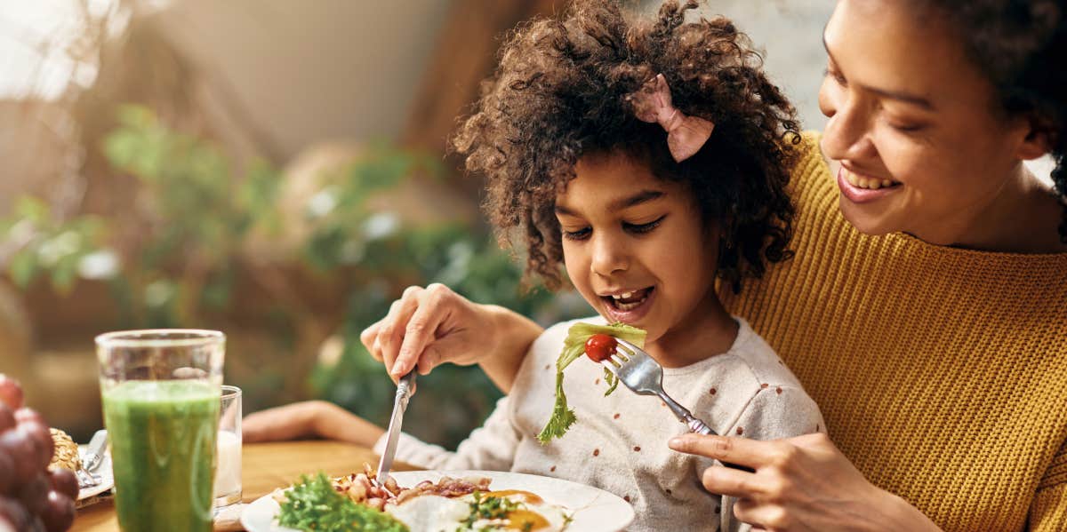 girl sitting on mother's lap while she is feeding her at dining table.