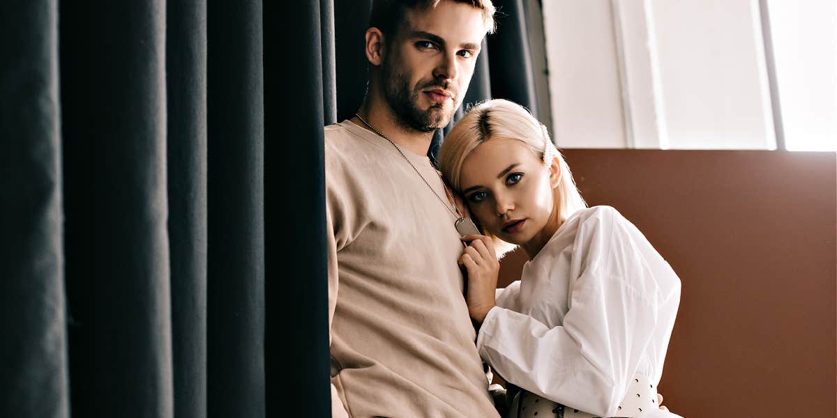 Brunette man with short beard and sweatshirt hugging well-dressed bleached blonde in fancy clothes - both looking serious 