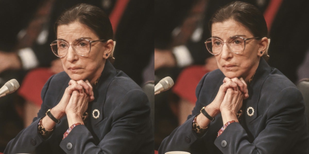 Justice Ruth Bader Ginsburg, in middle age, sits in front of a microphone wearing a grey suit, listening intently