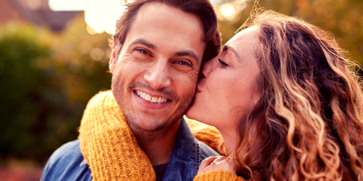 husband looking at camera smiling while wife kisses his cheek